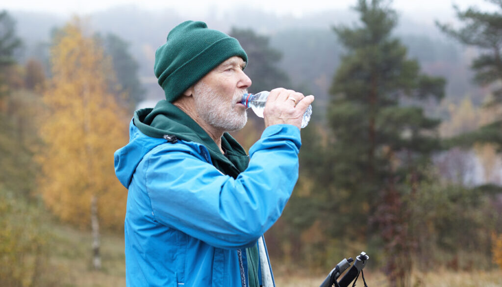 Sports, active lifestyle and refreshment concept. Autumn portrait of handsome sporty male pensioner with thick white beard refreshing himself after nordic walking, drinking water from plastic bottle