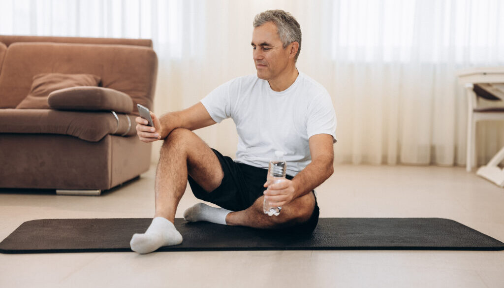 Put your phone down if you want a strong body! Procrastination during workout. Senior man sitting on yoga mat and texting on smartphone. Old man takes a rest after hard workout. krátky tréning na doma
