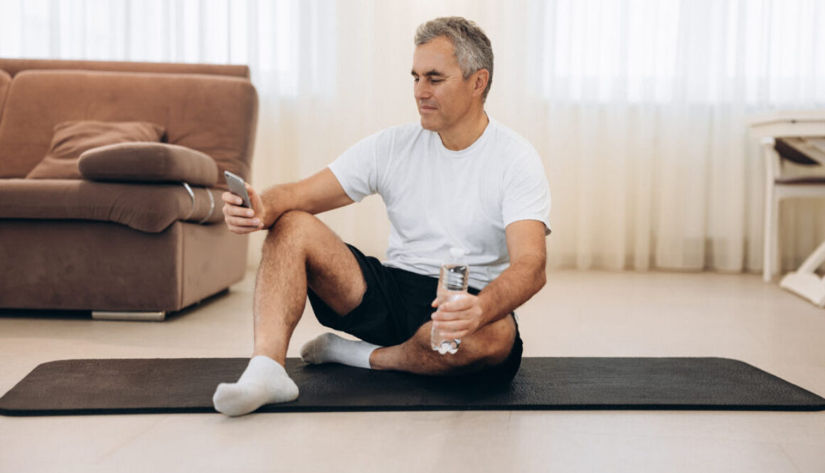 Put your phone down if you want a strong body! Procrastination during workout. Senior man sitting on yoga mat and texting on smartphone. Old man takes a rest after hard workout. krátky tréning na doma