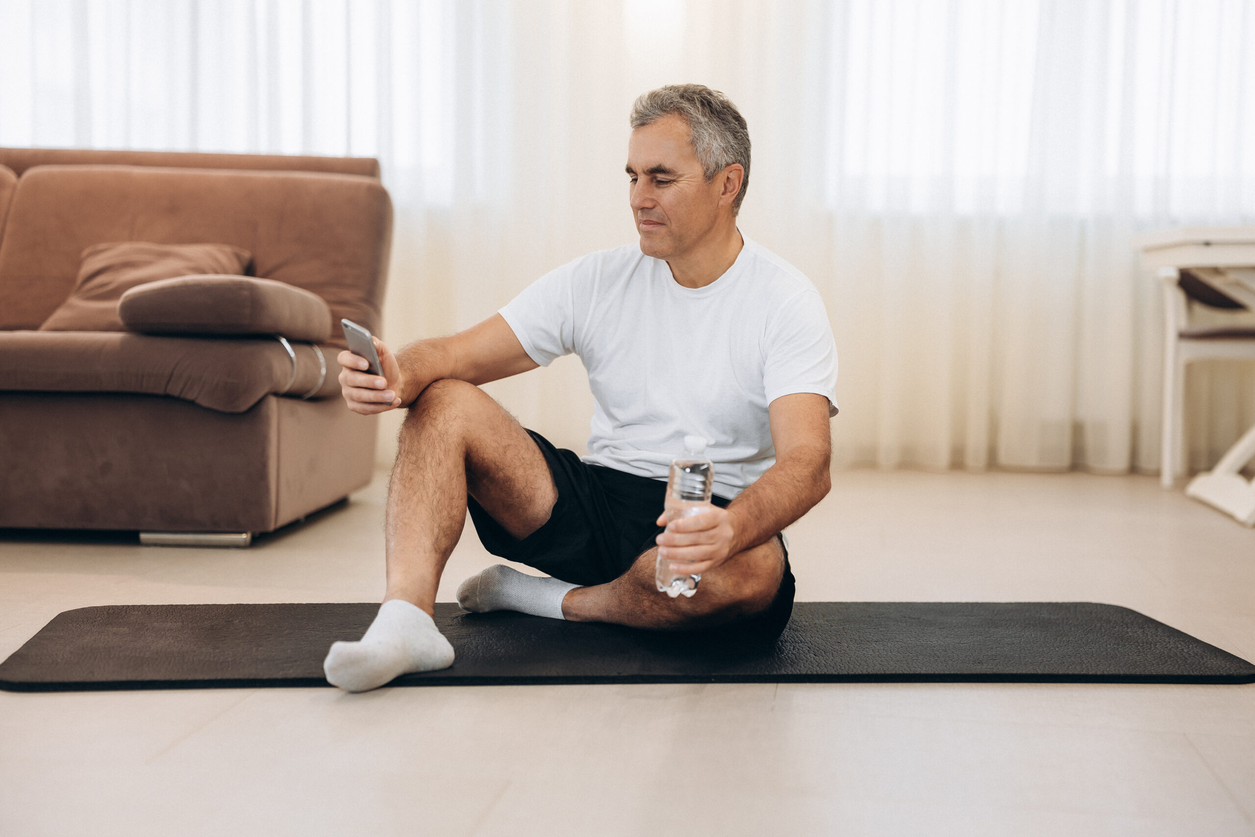 Put your phone down if you want a strong body! Procrastination during workout. Senior man sitting on yoga mat and texting on smartphone. Old man takes a rest after hard workout.